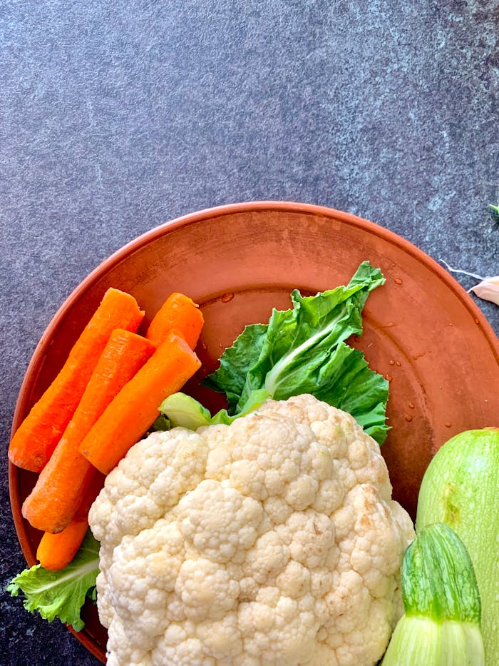 Top view of fresh carrots, cauliflower, and zucchini on a terracotta plate.
